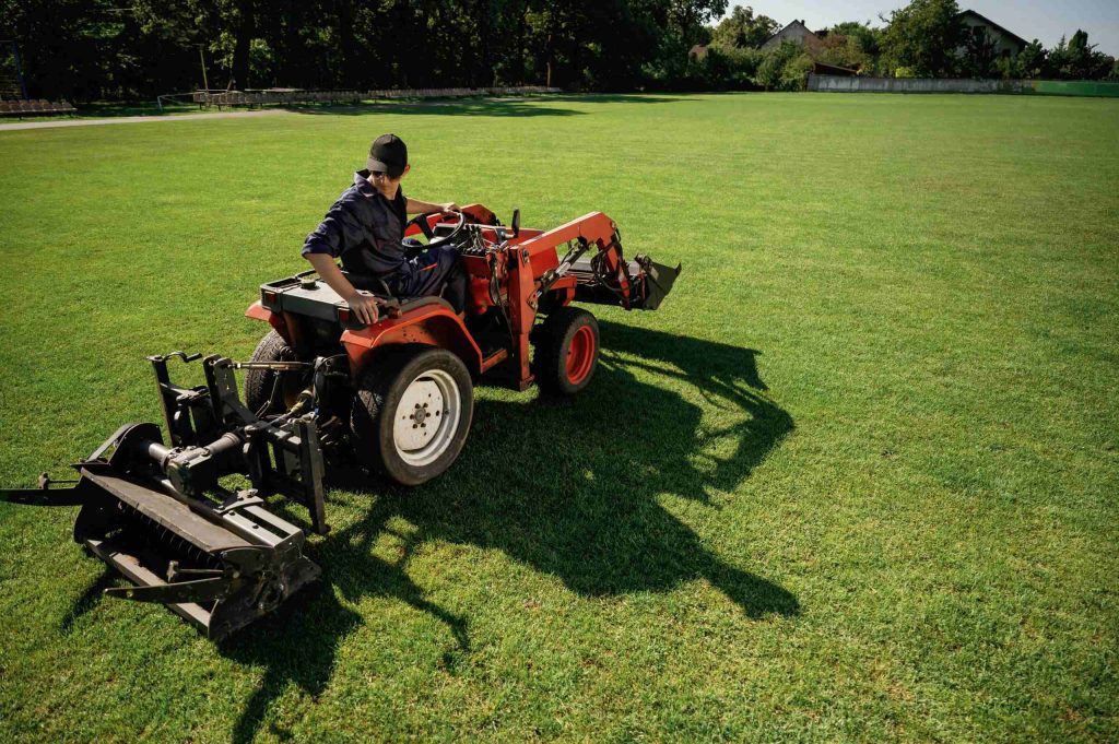 Commercial lawn care service worker operating a compact utility tractor on a vast, sunny green grass stadium field.