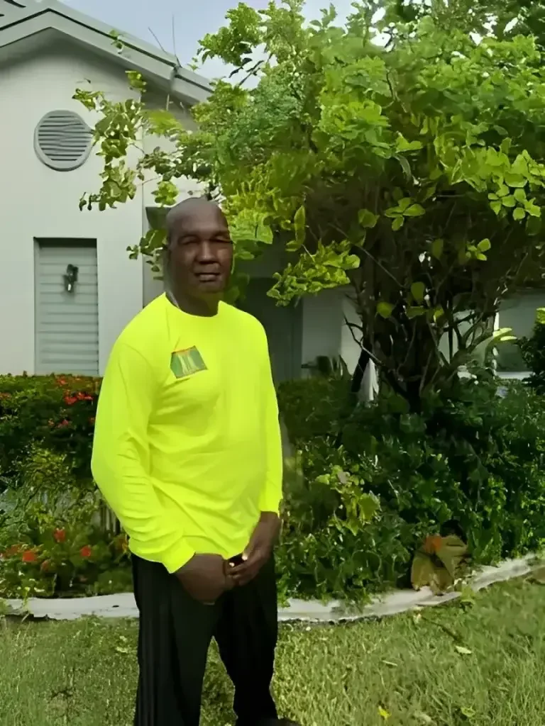 Professional lawn care contractor in a neon yellow shirt standing by a neatly trimmed tree and lush garden landscape.
