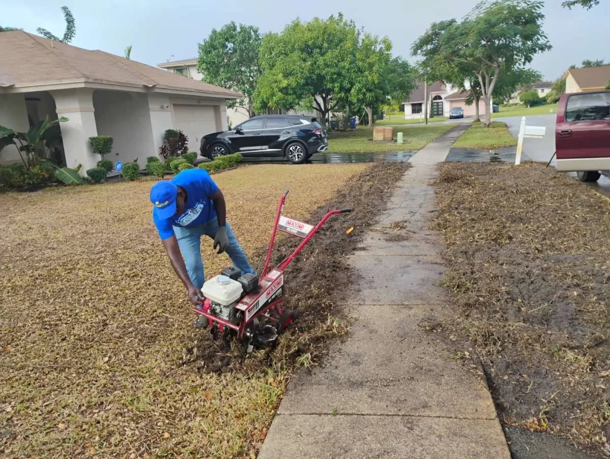 A man in a blue cap tilling the front yard grass with a red rototiller machine next to a concrete neighbourhood walkwa